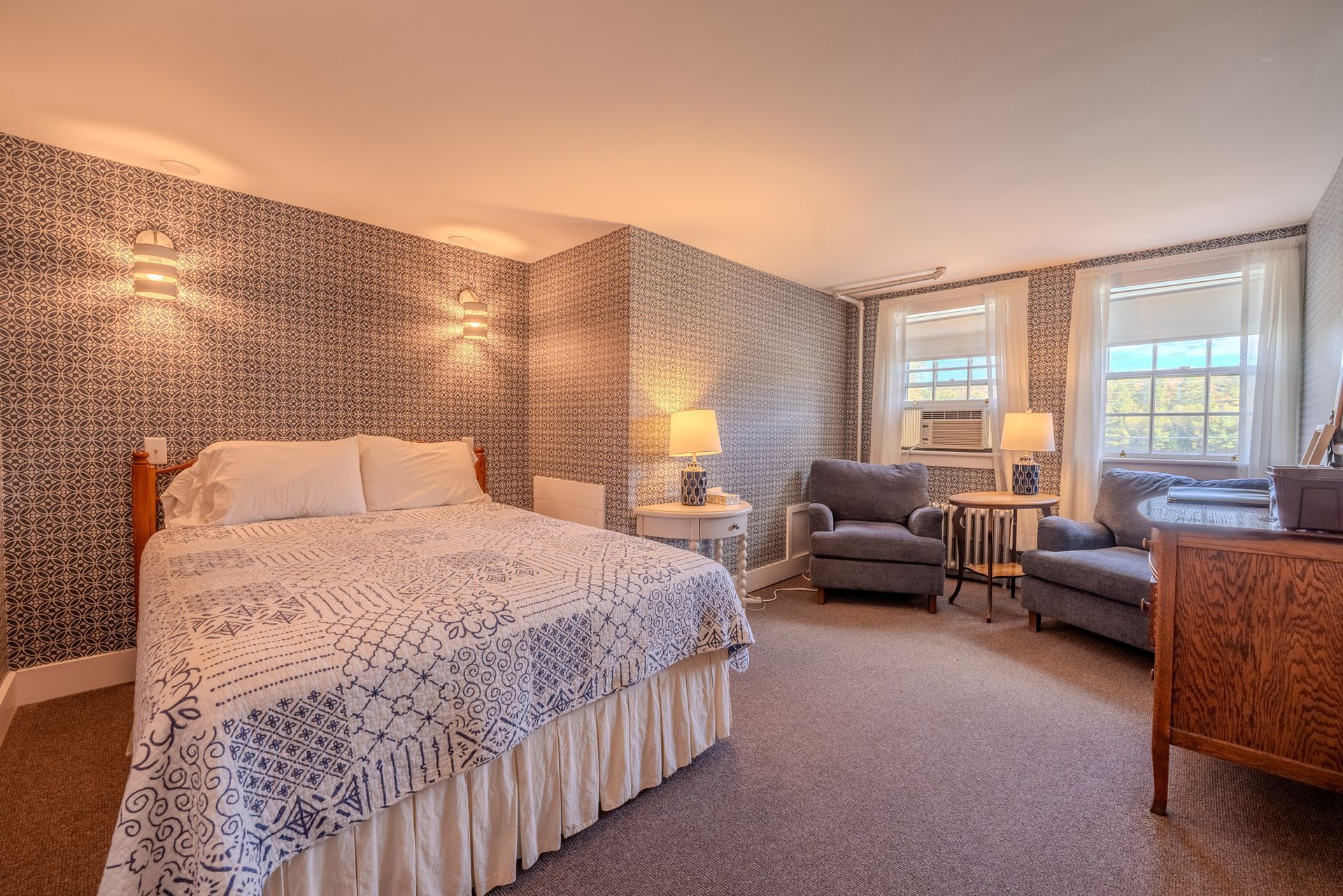 Bedroom with patterned wallpaper, bed, two armchairs, lamps, and windows.