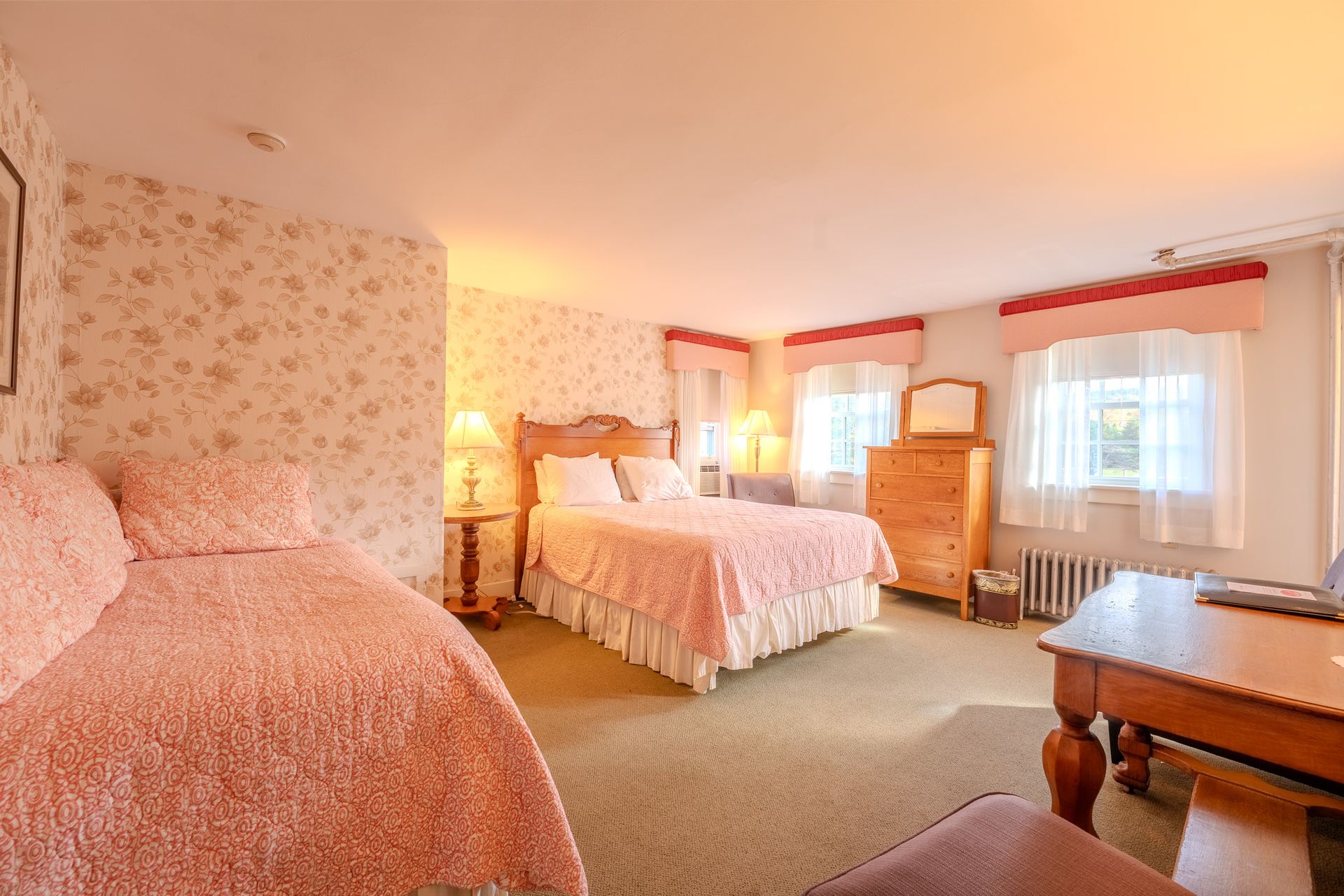 A bedroom with floral wallpaper, featuring a bed, a pull-out bed, dresser, and desk; illuminated by warm lighting.