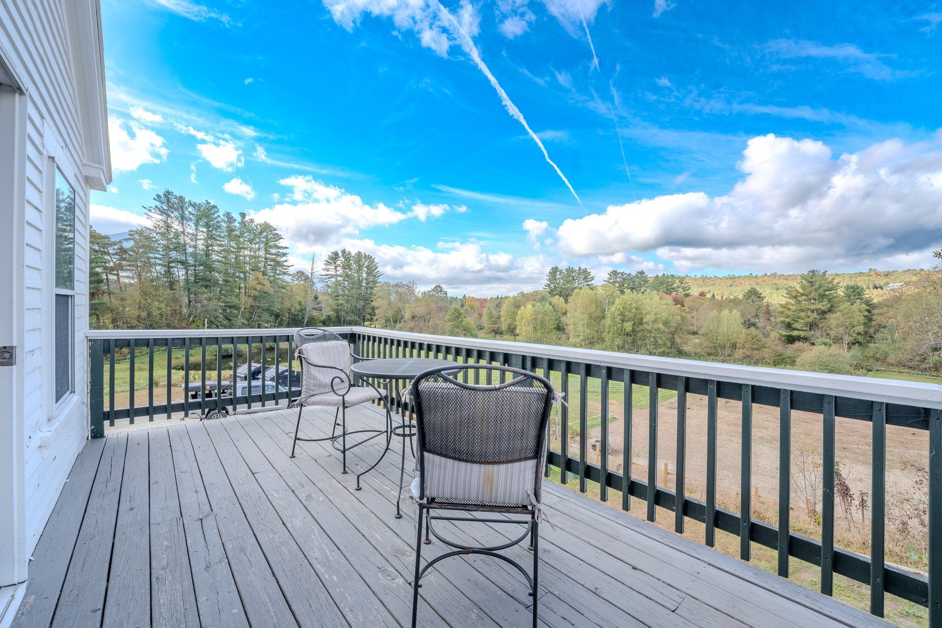 Wooden deck overlooking a rural landscape with two chairs and a small table under a bright blue sky.