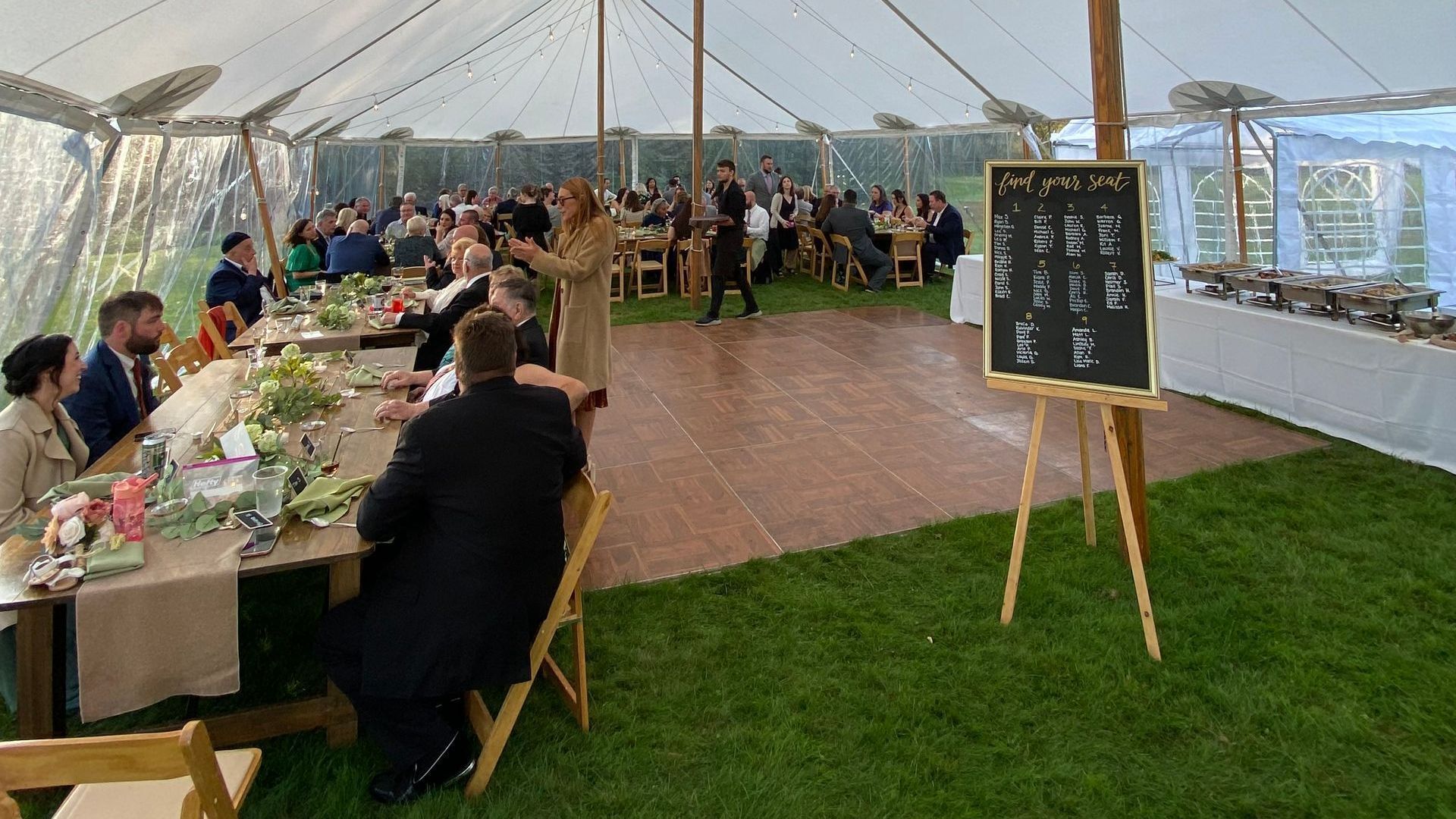 Wedding reception tent with guests, tables, a dance floor, and a menu board on a wooden easel.