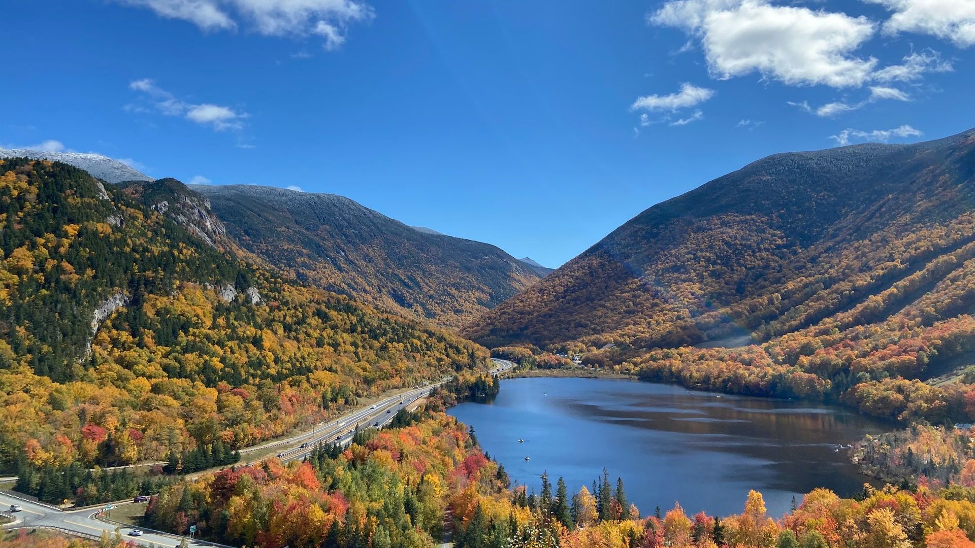Fall foliage surrounds a lake nestled between mountains under a blue sky.
