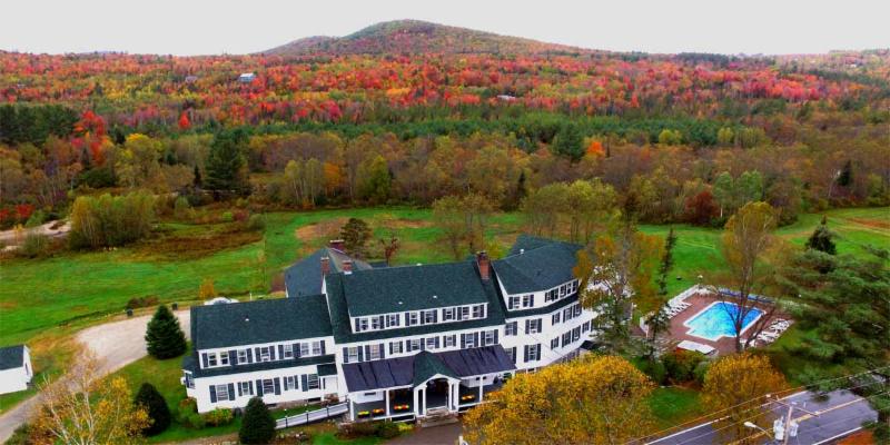 A large white building with green roof sits among fall foliage; mountains in the background.