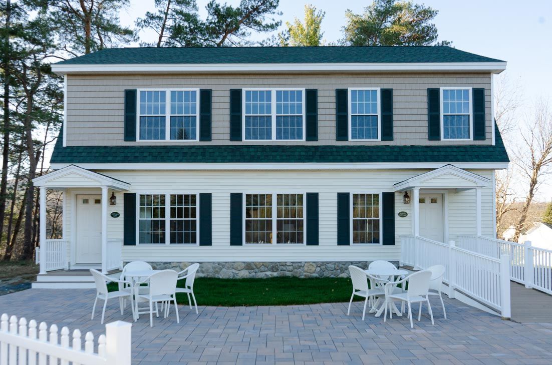 Two-story building with white siding, green roof, and shutters. Paved patio with tables and chairs.