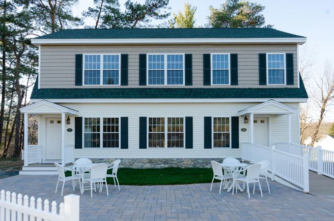 Two-story duplex with white and tan facade, green roof and shutters, tables and chairs on brick patio.