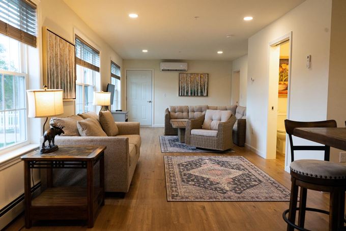 Cozy living room with a tan sofa, armchair, rug, and side tables. Includes a small kitchen bar and bathroom doorway.