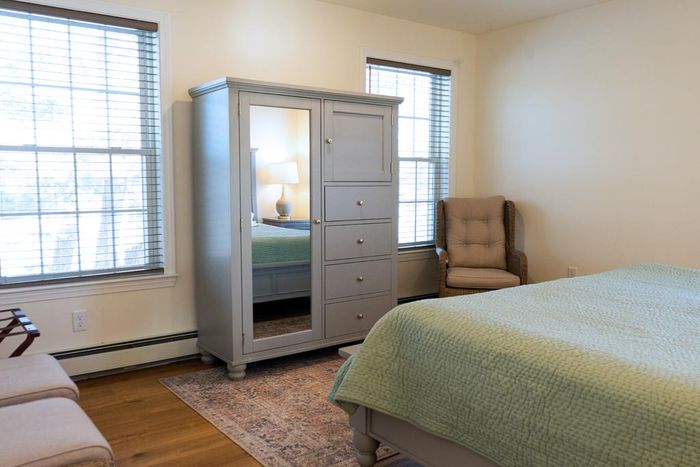 Bedroom with a gray armoire, bed with green quilt, windows, and a chair.