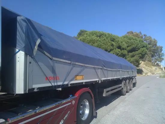 Un grande camion con rimorchio, coperto da un telone blu, è parcheggiato a bordo strada sotto un cielo sereno.