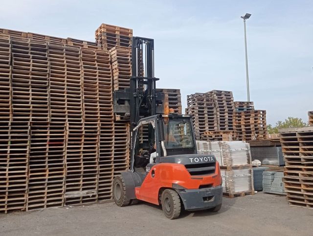 Un carrello elevatore arancione in un deposito di legname, carico di pallet e casse di legno.