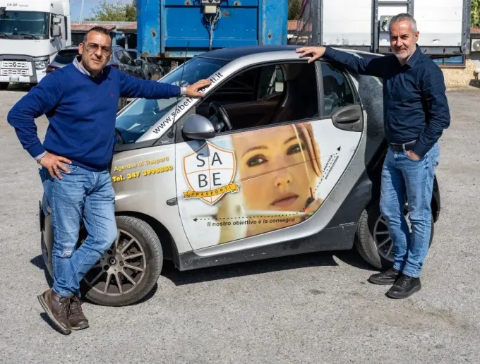 Due uomini sono in piedi accanto a una piccola auto bianca con una grande grafica raffigurante un volto, parcheggiata all'aperto in un parcheggio.