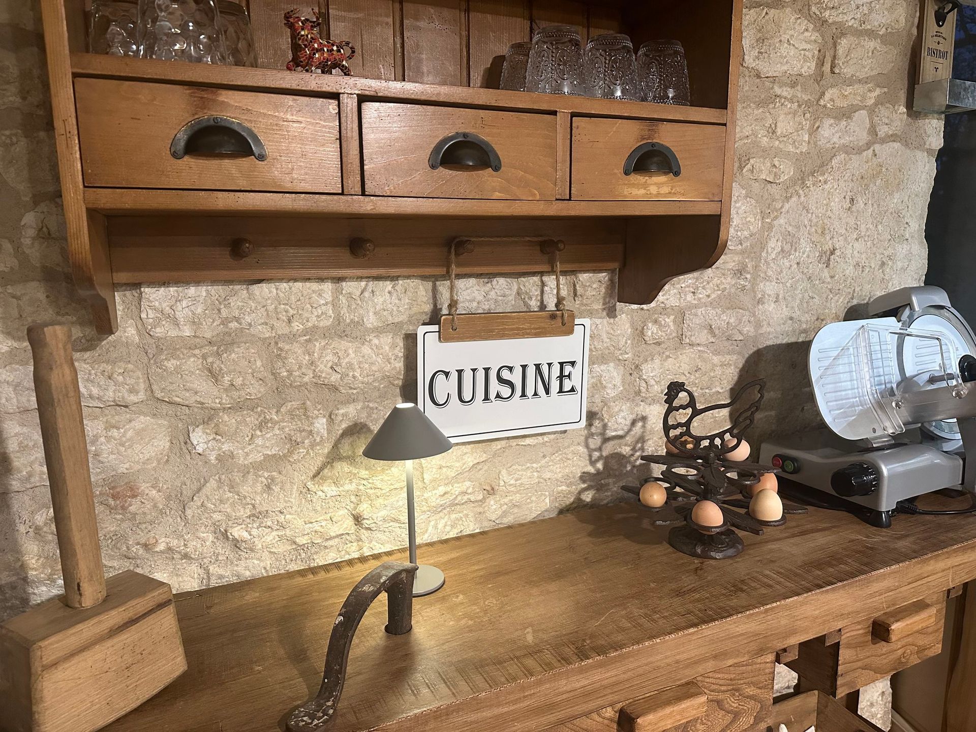 Plan de travail de cuisine avec étagères en bois, tiroirs et panneau « CUISINE ». Comprend une lampe, des œufs et une trancheuse.