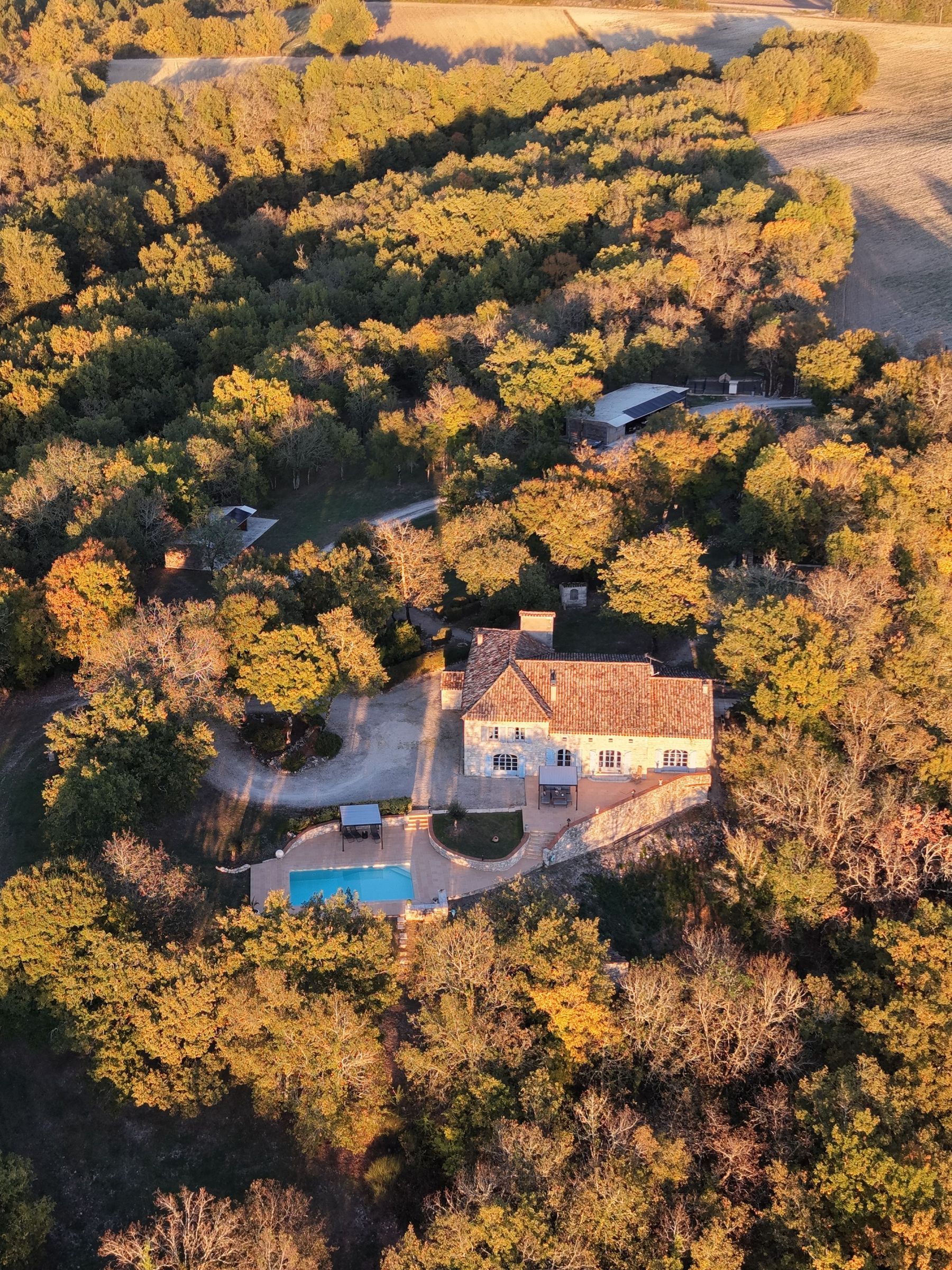 Vue aérienne d'une maison avec piscine entourée d'arbres au coucher du soleil.