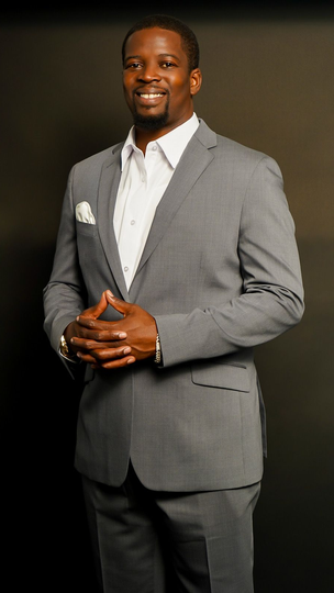 Man in a light gray suit standing and smiling against a dark studio background