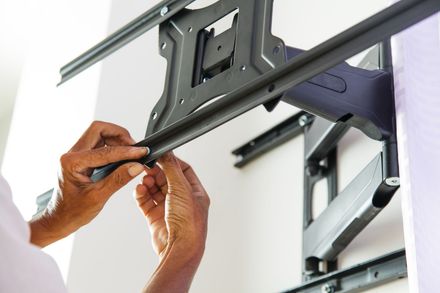 Hands attaching a black TV mount to a wall bracket near a ladder