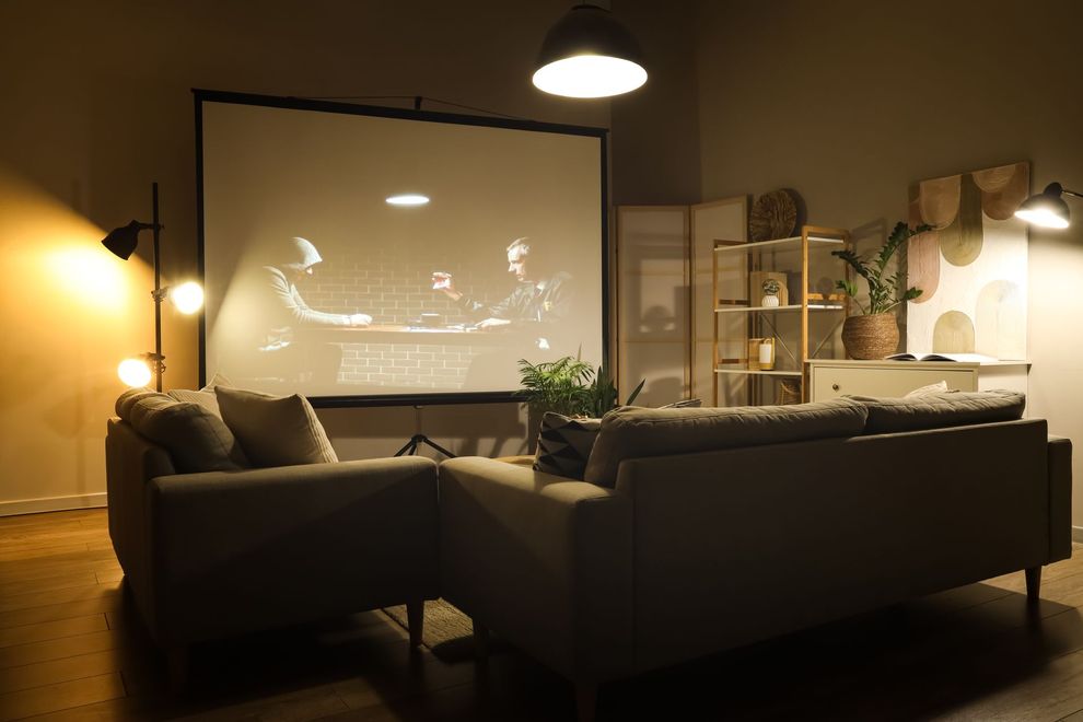 Cozy living room with sofas facing a projected movie screen, warm lamp lighting, and plants.