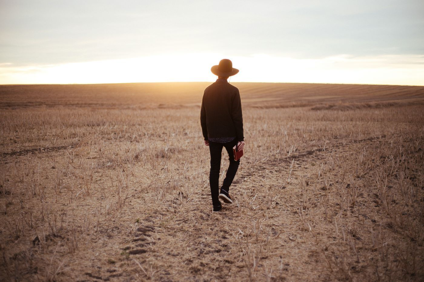 A person in a hat walking across a barren field toward a bright sunset.
