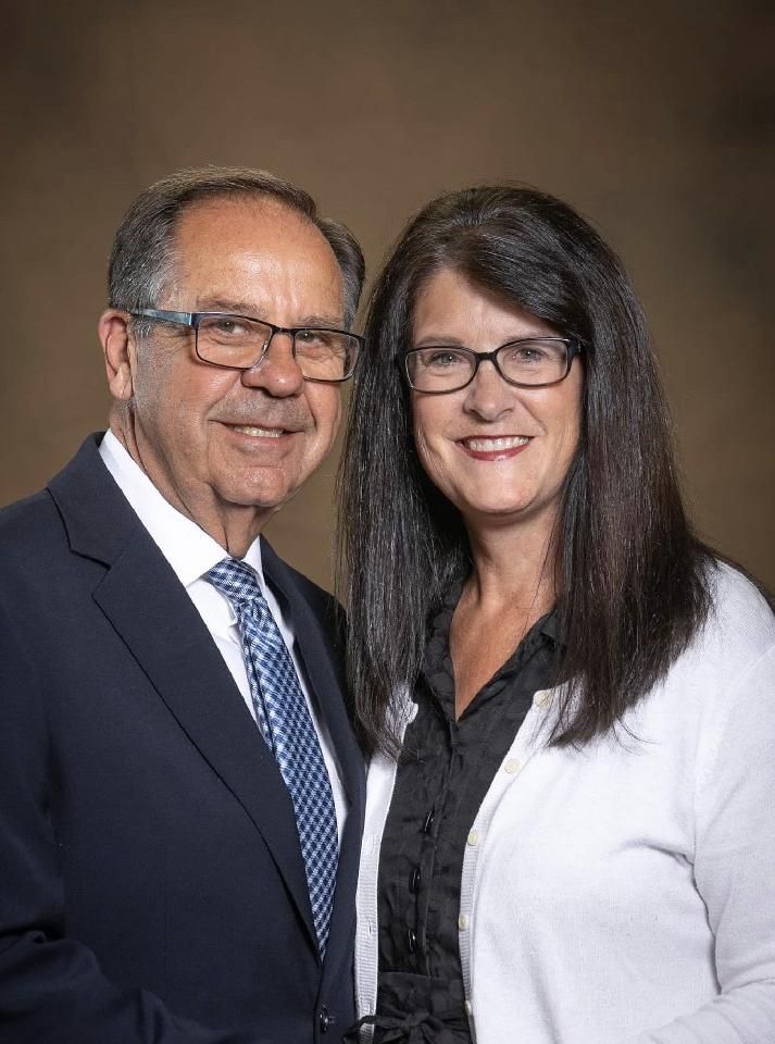Smiling couple posing together against a plain backdrop, wearing business attire and glasses.