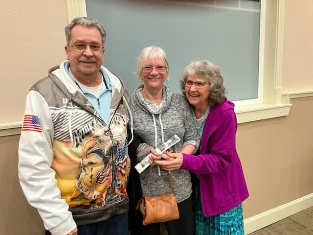 Three people smiling and standing together indoors by a window.