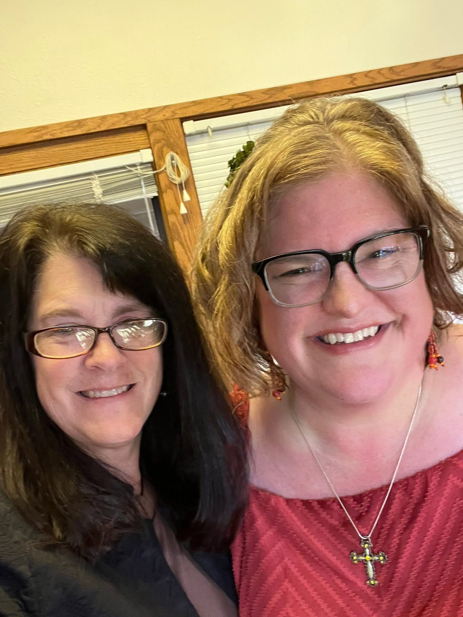 Two smiling women with glasses posing indoors against a wooden wall.