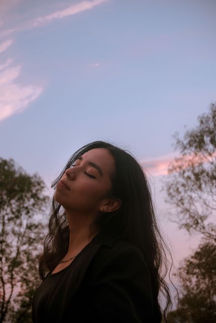 Mujer con los ojos cerrados, vestida con una blusa negra, de pie al aire libre bajo un cielo rosado al atardecer.