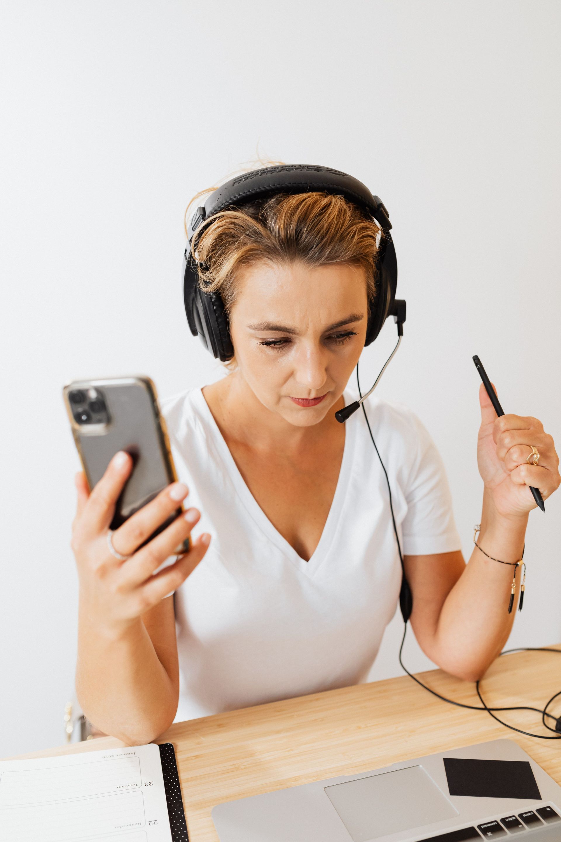 Persona con auriculares puestos, usando un teléfono inteligente mientras sostiene un bolígrafo en un escritorio con una computadora portátil.