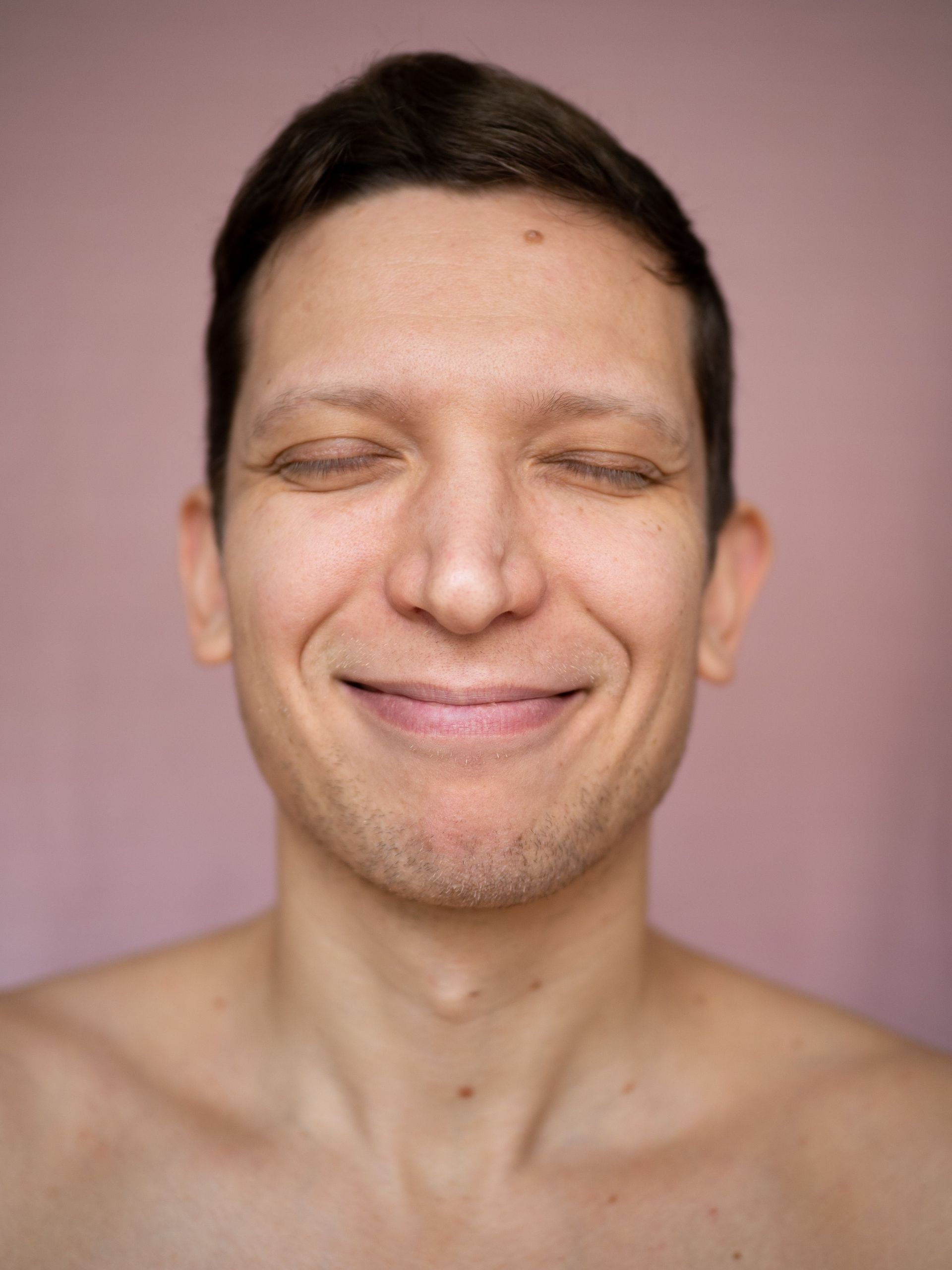 Retrato de una persona sonriente con los ojos cerrados sobre un fondo rosa.
