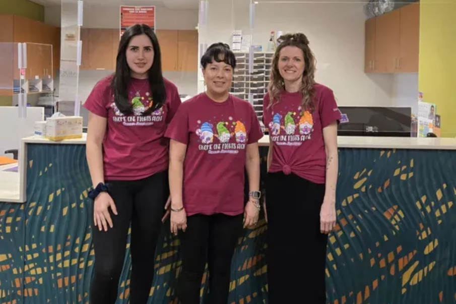 Three people stand behind a counter. They wear maroon shirts with gnome design, smiling. Interior setting.