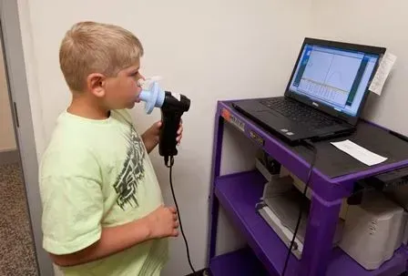 Boy exhales into a device connected to a laptop on a cart; medical test in progress.