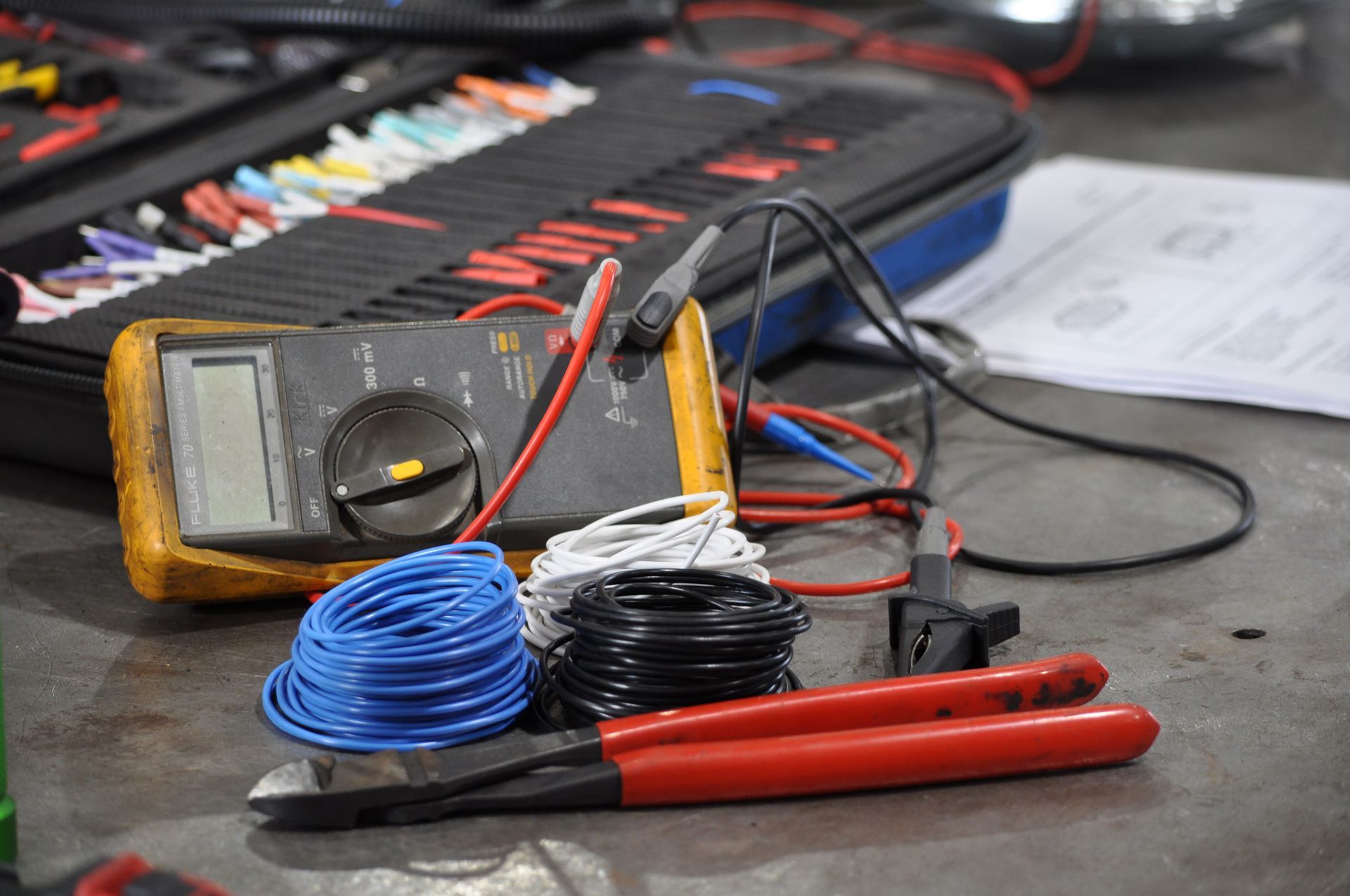 Analog multimeter with red and black test leads on a workbench, beside blue wire and red tools