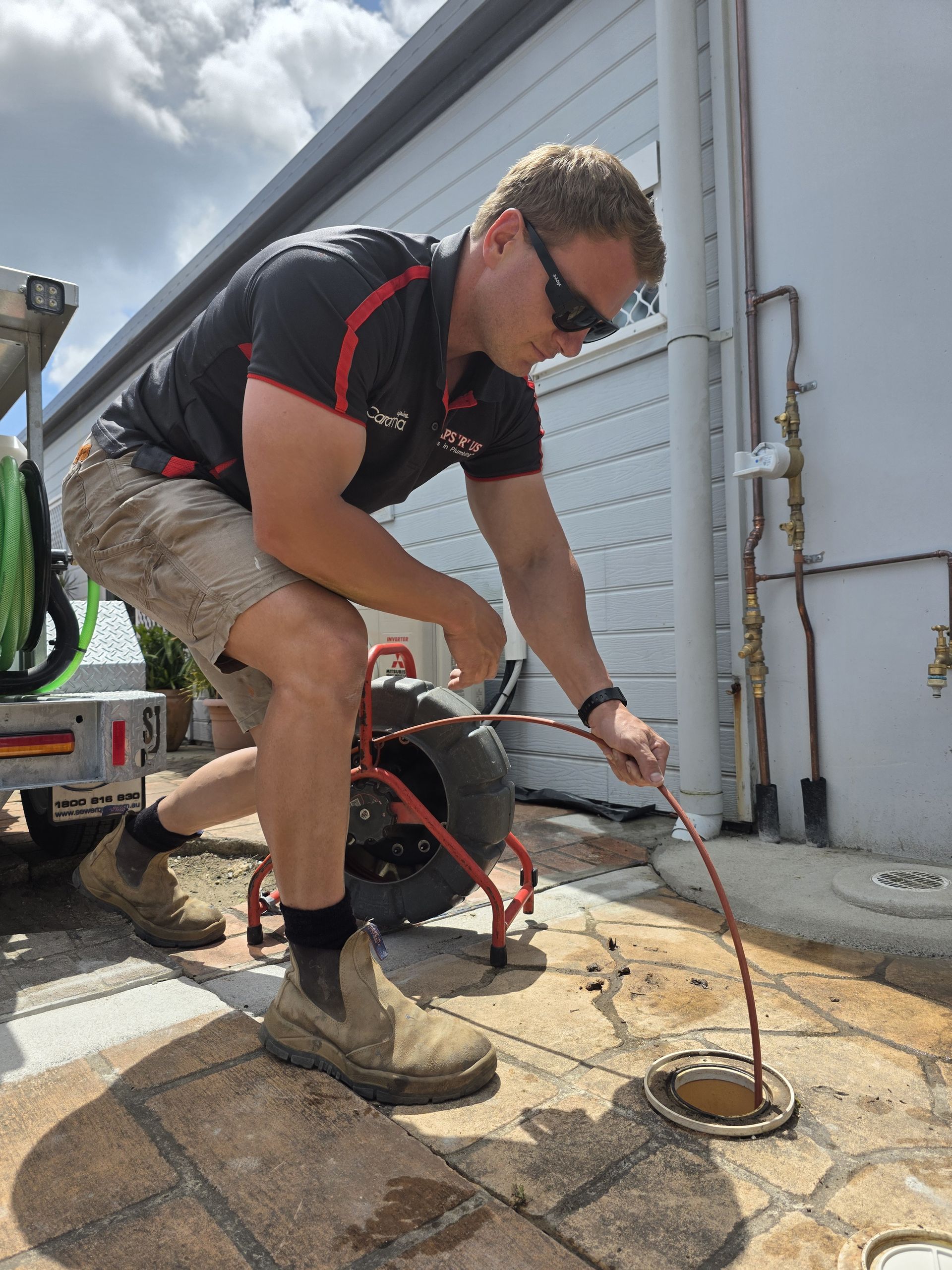 Worker Crouches by A Wall Using a Tool on A Metal Drain in An Outdoor Service Area — Taps 'R' Us in Kunda Park, QLD