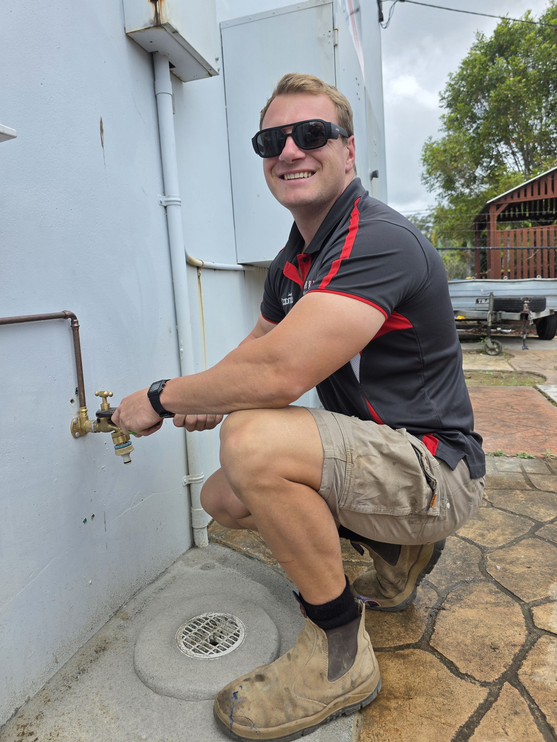 Man kneeling by an outdoor faucet, turning the valve, wearing sunglasses, work shirt, shorts, and boots. — Taps 'R' Us in Caloundra, QLD