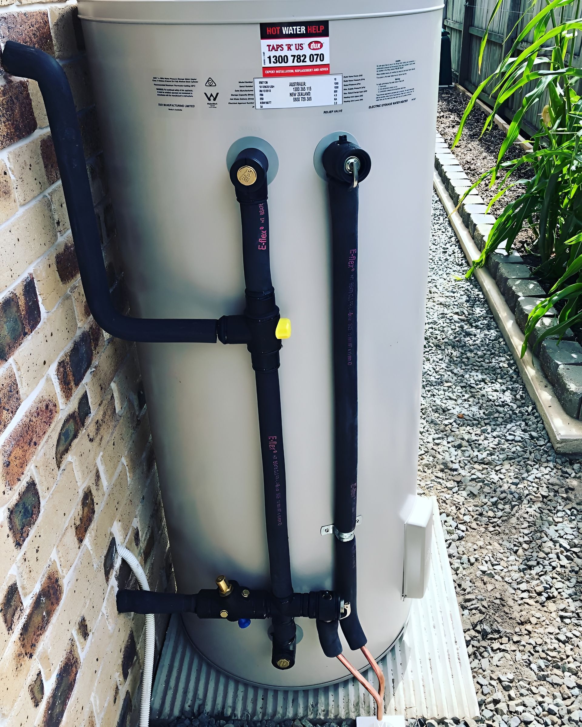 A cylindrical water heater with black pipes against a brick wall and a garden. — Taps 'R' Us in Kunda Park, QLD