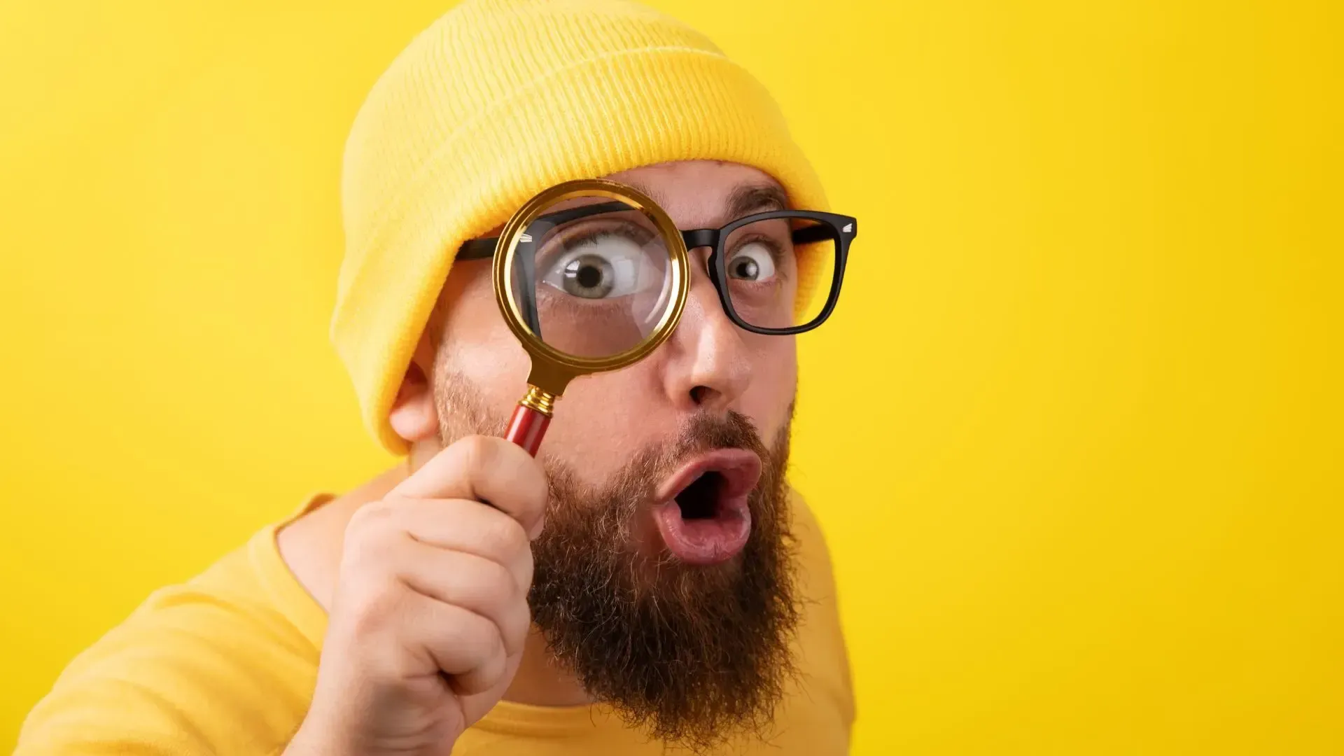 Man with yellow hat and shirt, glasses, and beard peers through magnifying glass, mouth open, against yellow background.