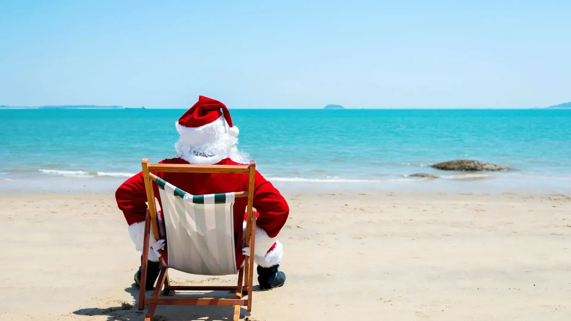 Santa Claus relaxing on a beach, facing the ocean. He is wearing his red suit and hat, sitting in a folding chair.