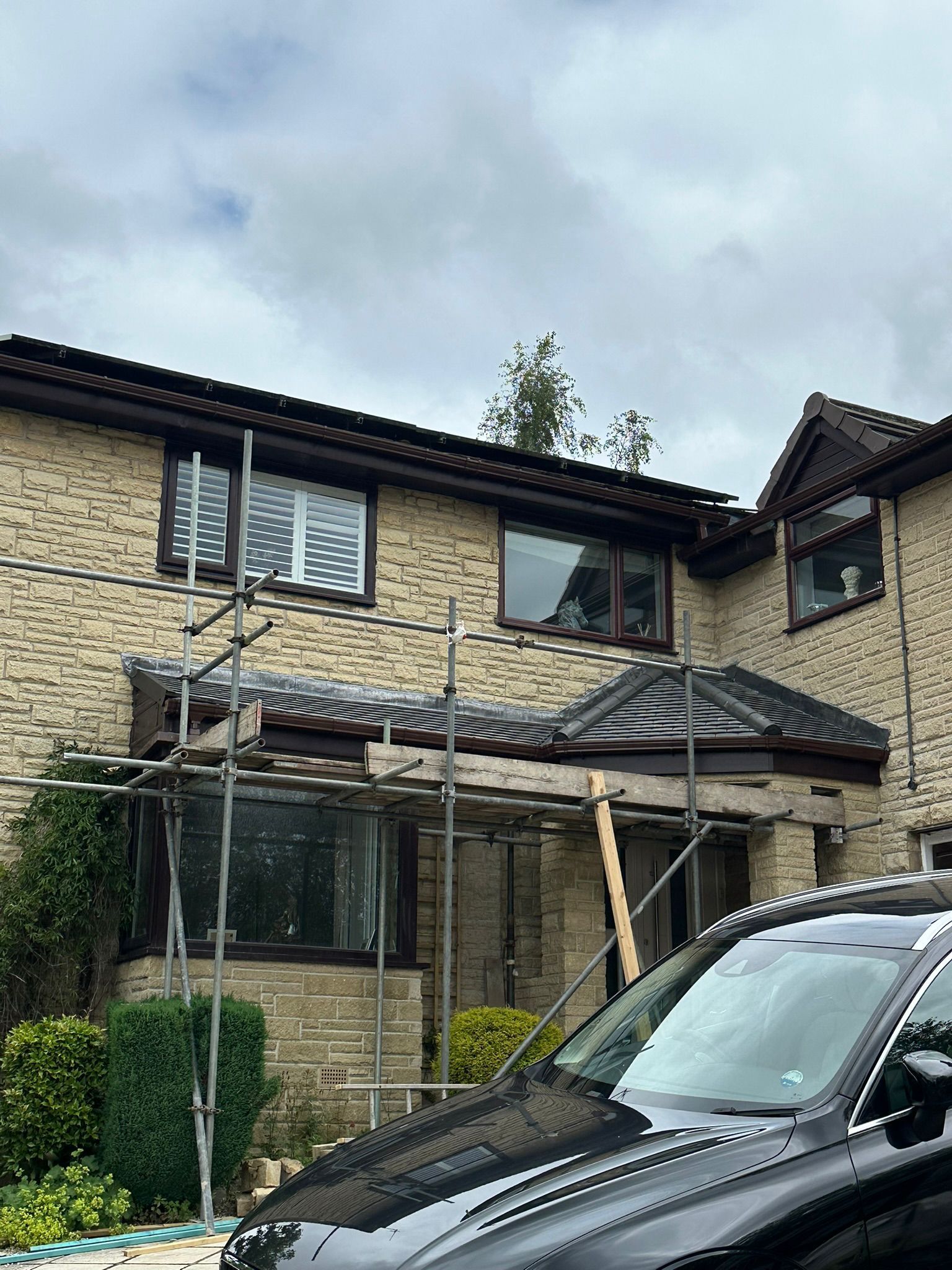 Brick house with damaged roof and scaffolding under cloudy sky, black car in foreground