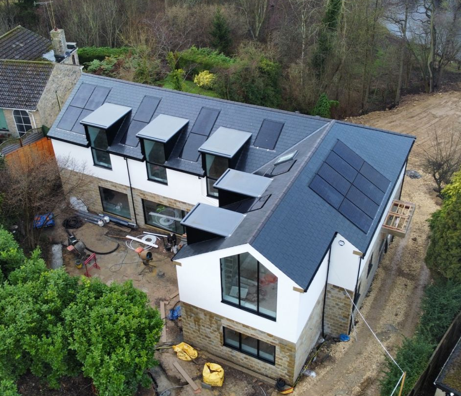 Aerial view of a white house with dark roof solar panels and dormers, surrounded by trees and a yard.