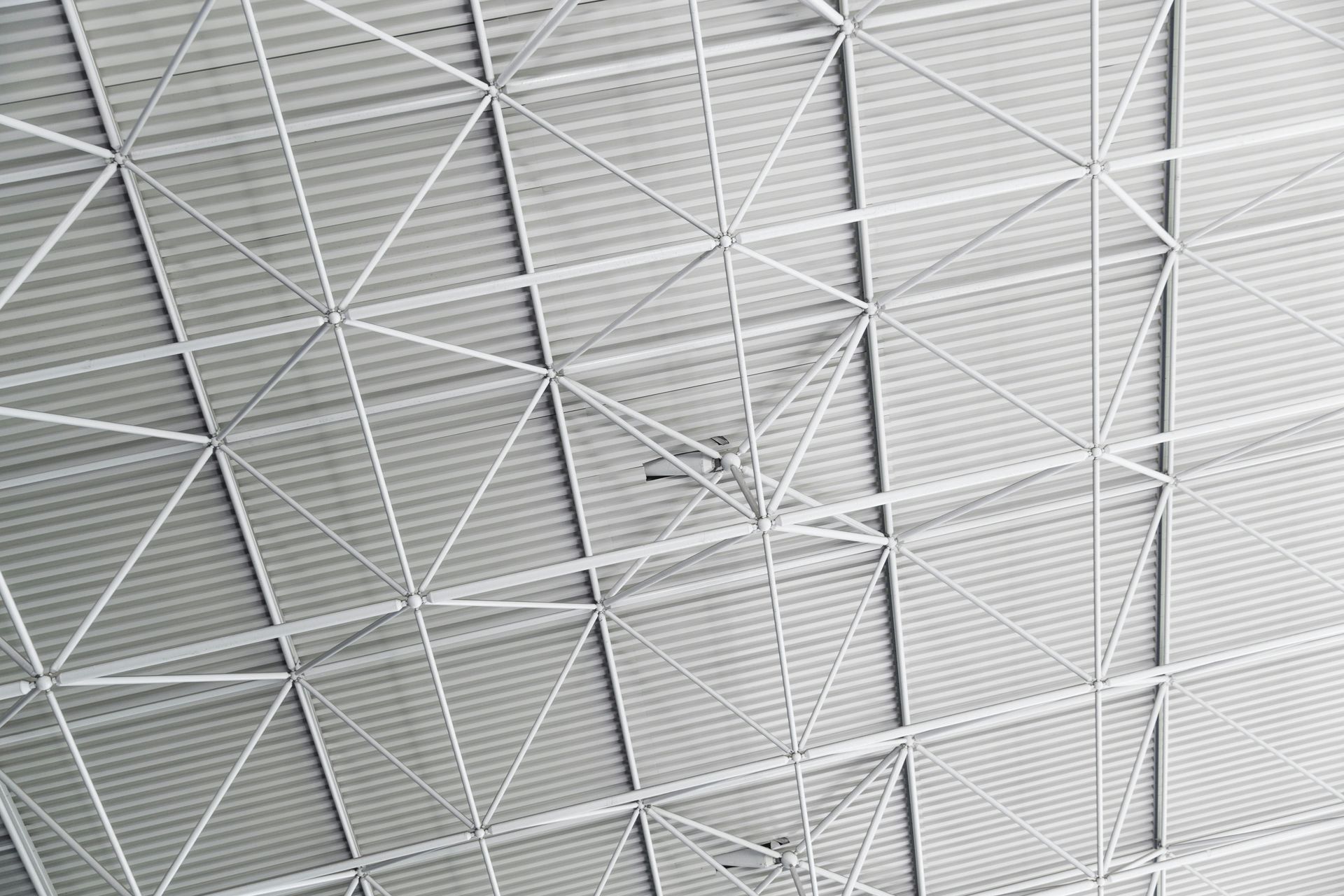 An industrial metal ceiling with a repeating pattern of white trusses and support beams against a corrugated roof.