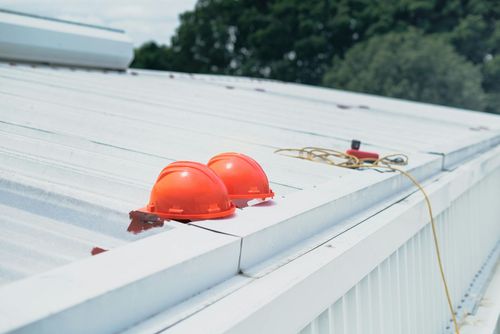 Two orange hard hats and a coiled yellow safety rope sit on a white metal industrial roof.