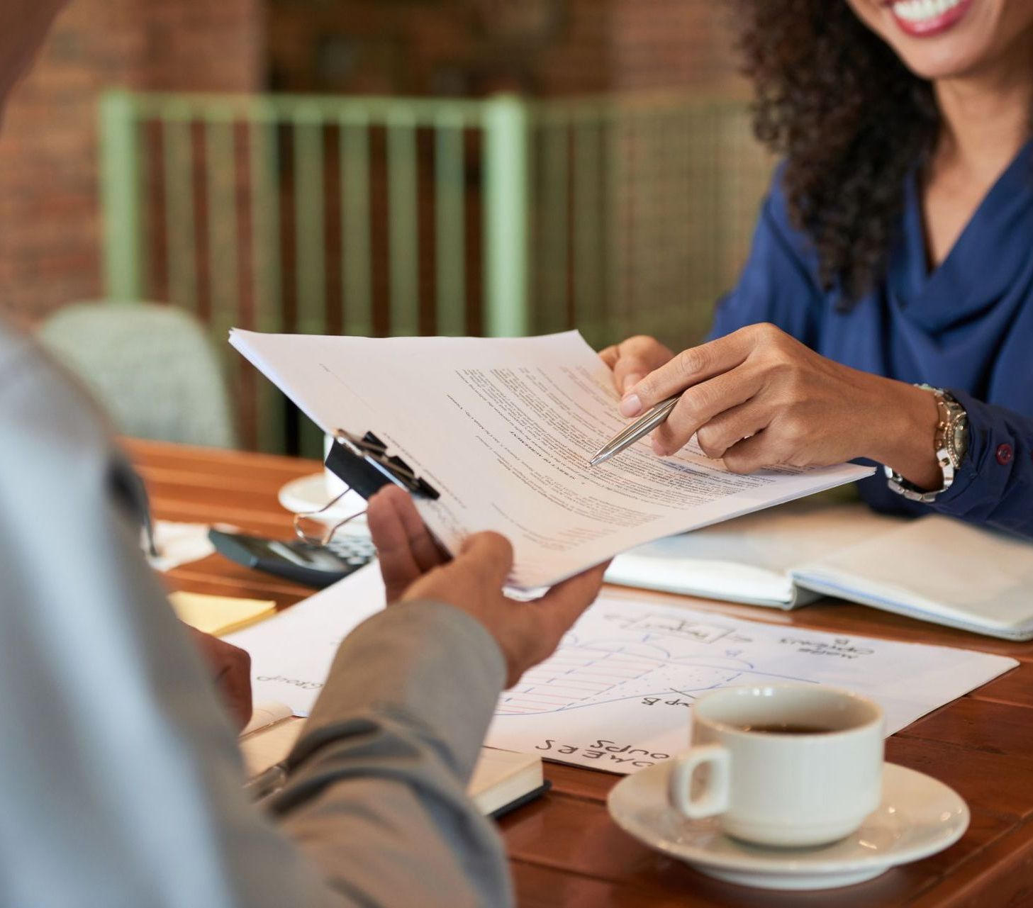 A woman is holding a piece of paper and a pen while sitting at a table with a cup of coffee.