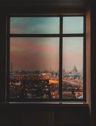 City skyline at dusk seen through a window, with warm orange lights and a pink-blue sky.