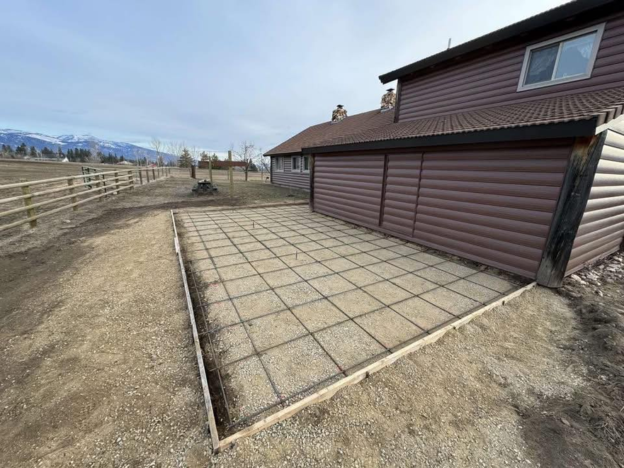 A rectangular concrete slab forms a patio next to a log-sided house, featuring a dark metal rebar grid ready for pouring.