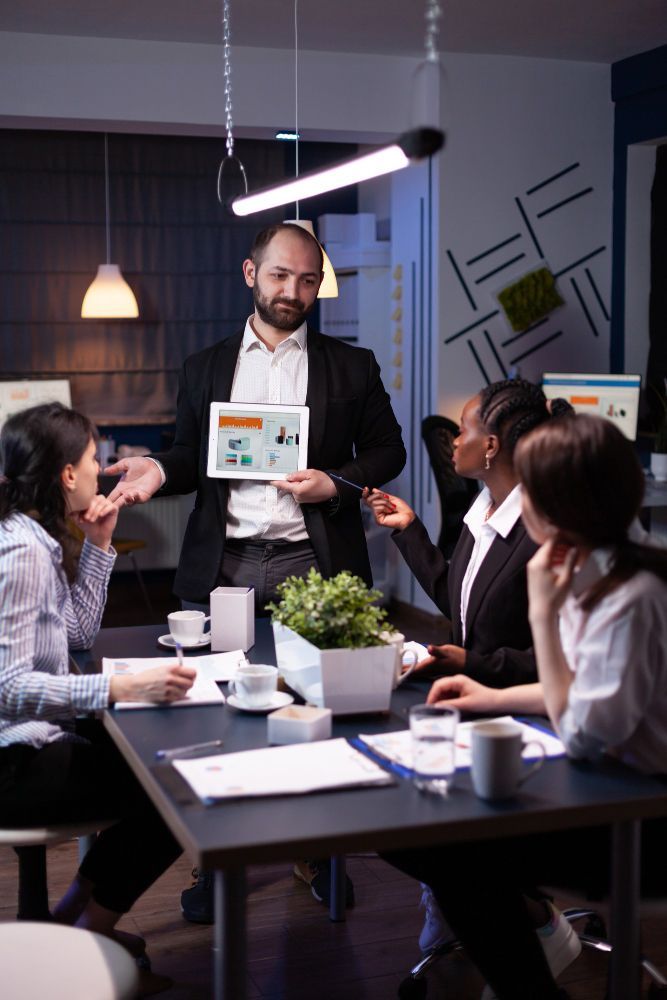 A professional team holds a late-night meeting around a table, listening as a person presents data on a digital tablet.
