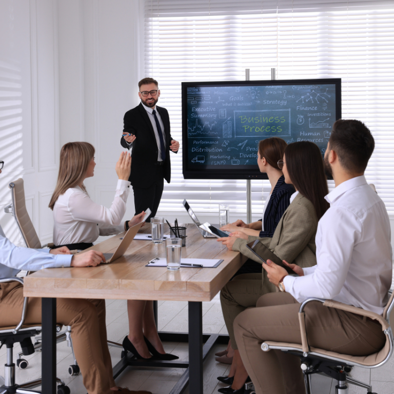 A professional presenter stands before a screen while colleagues sit at a table, one raising a hand during a meeting.