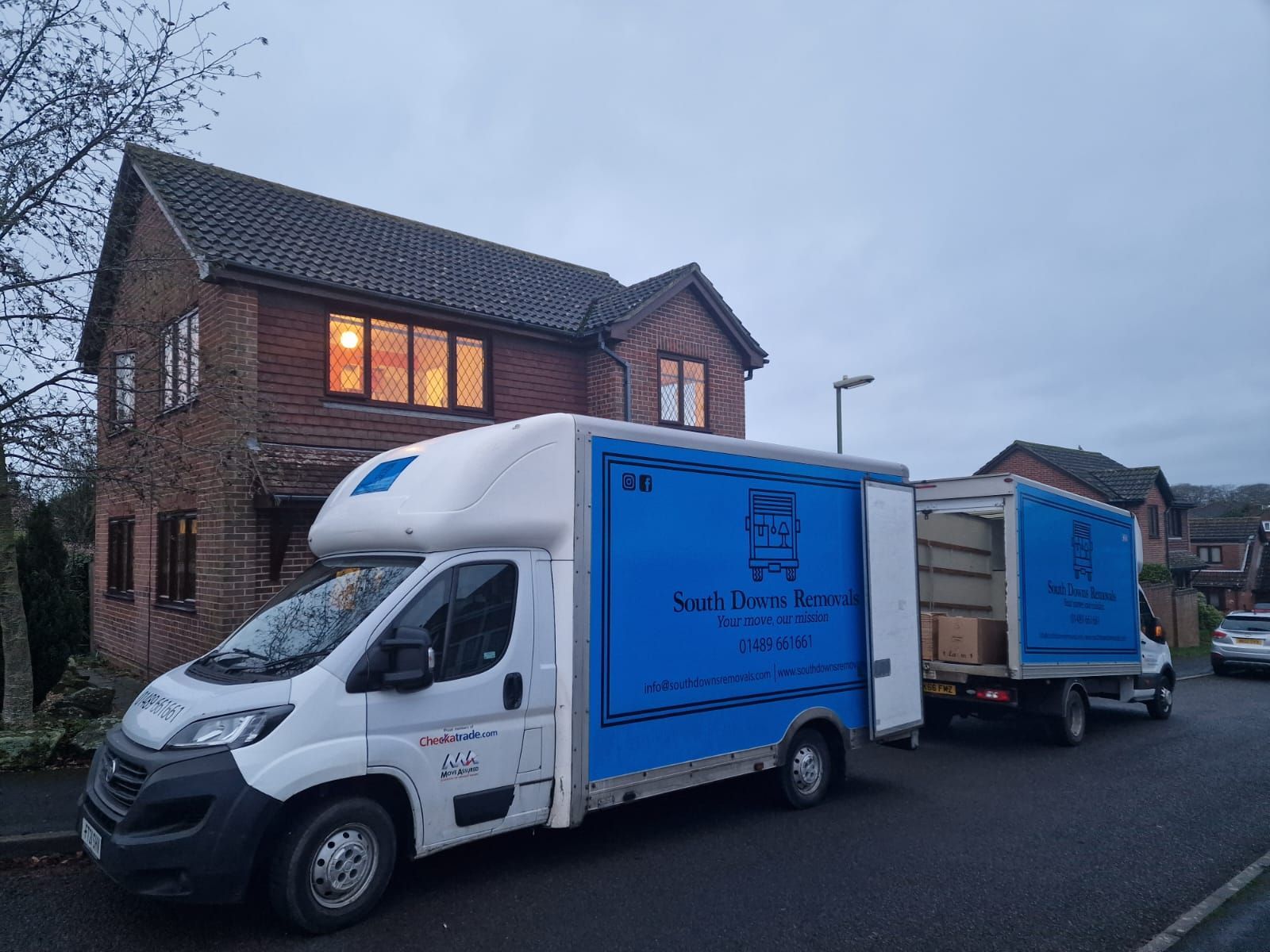 Two blue and white moving trucks parked in front of a house, boxes visible.