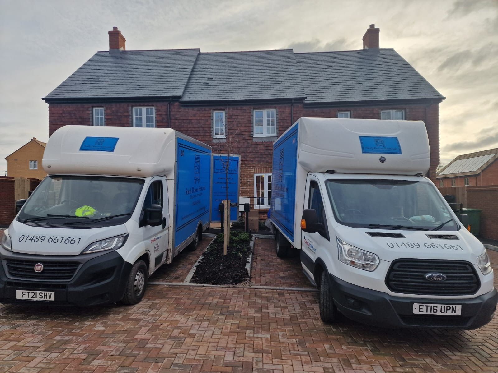 Two white and blue moving vans parked in front of a brick house.