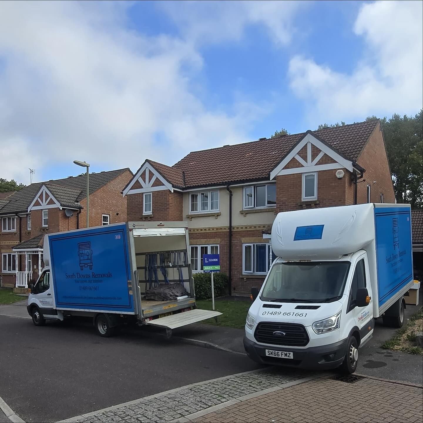 Two blue moving trucks parked in front of a residential home on a sunny day.