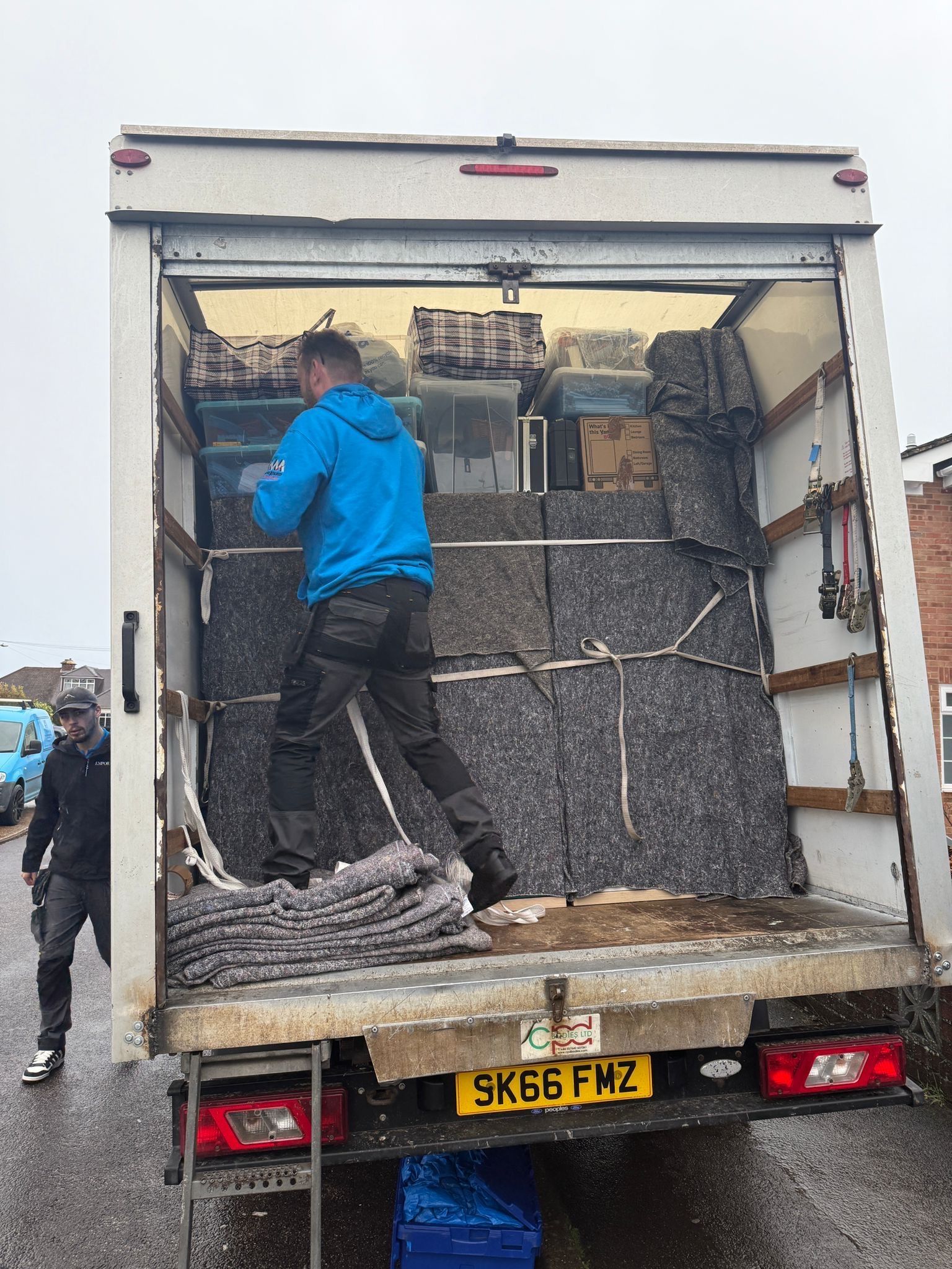 Two people unloading a van. One stands inside, the other walks. Van is full of items.