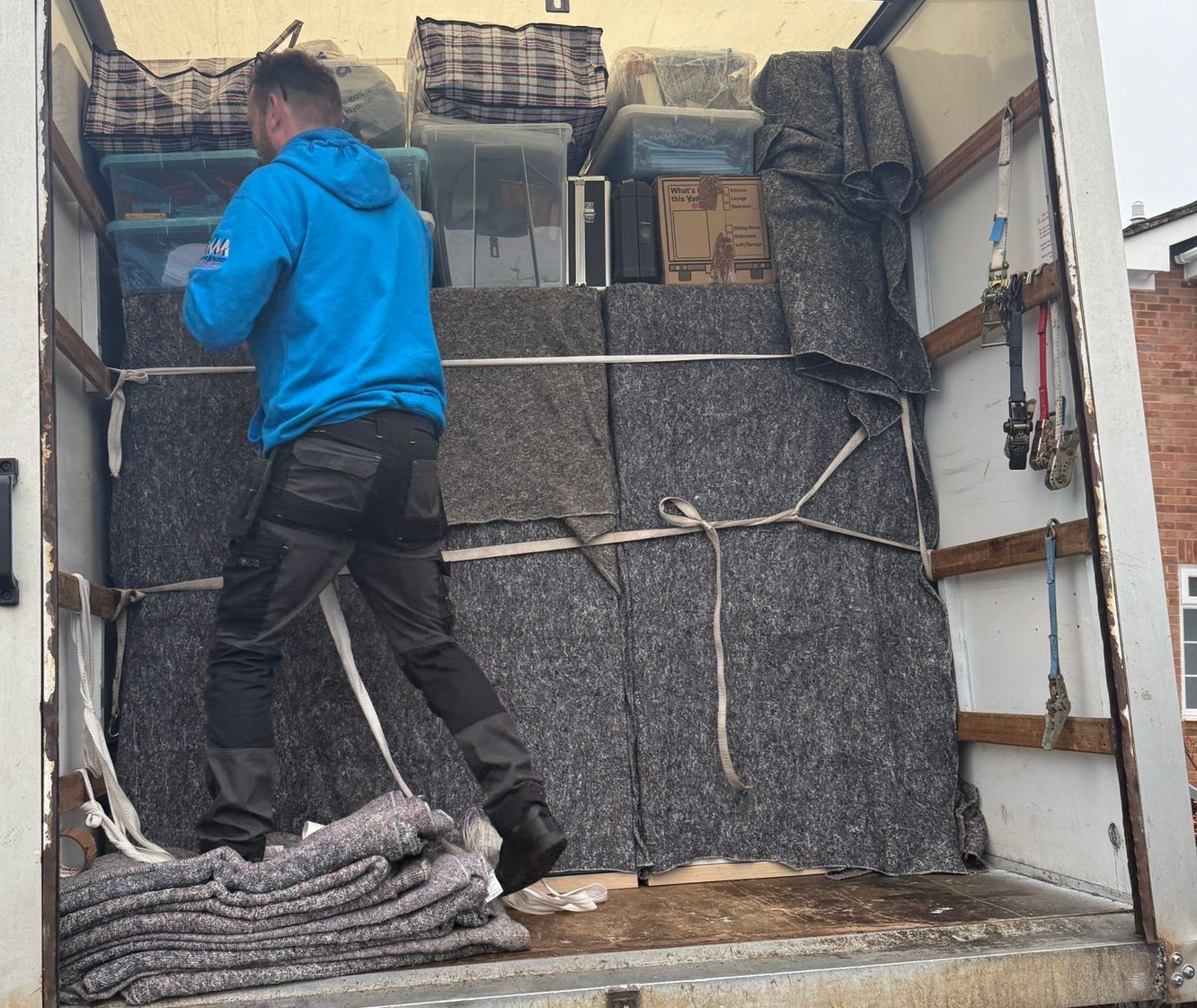 Person in blue jacket loads a moving truck with boxes, secured with blankets and rope.