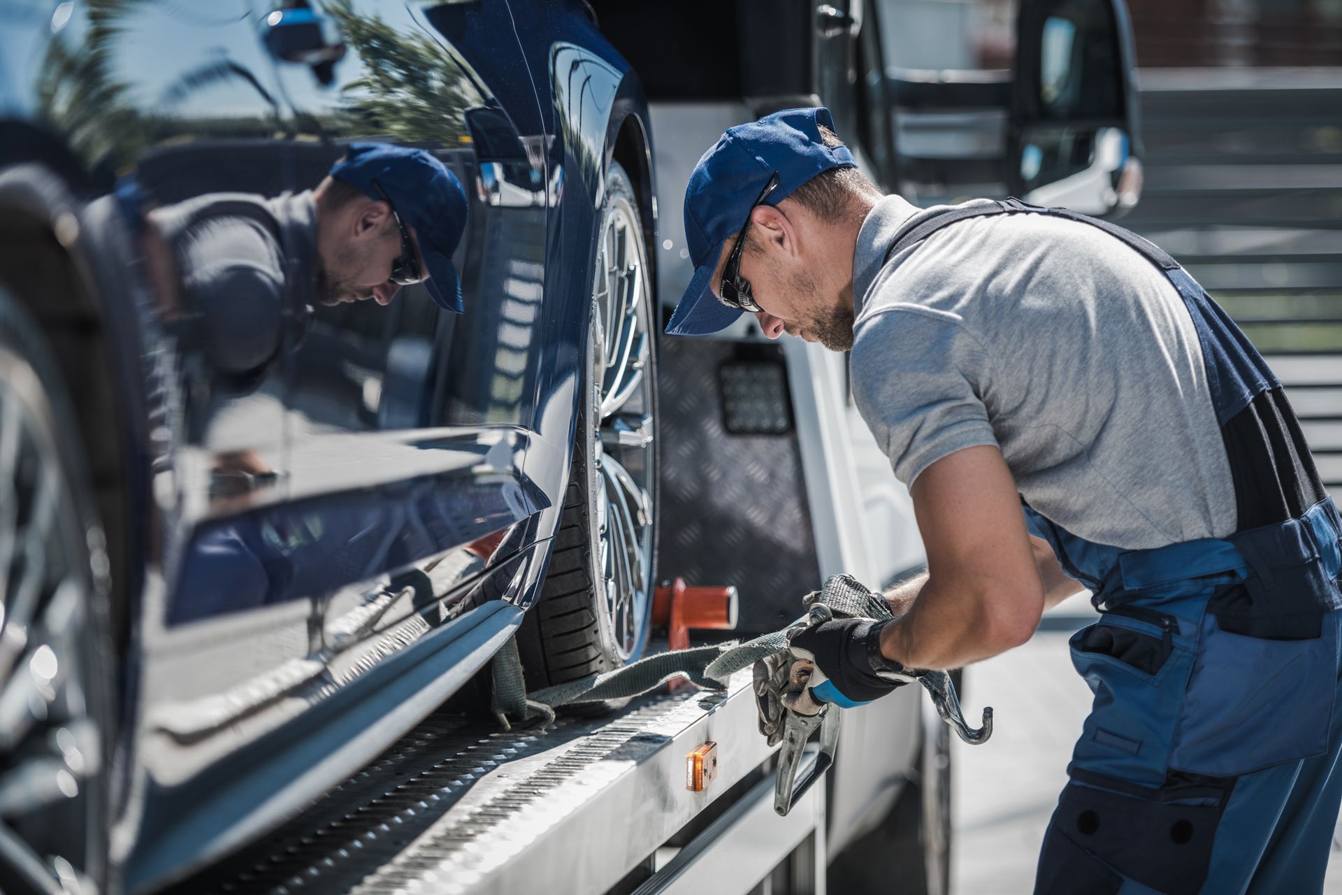 A professional in a blue cap and work overalls secures a dark car to the bed of a tow truck.
