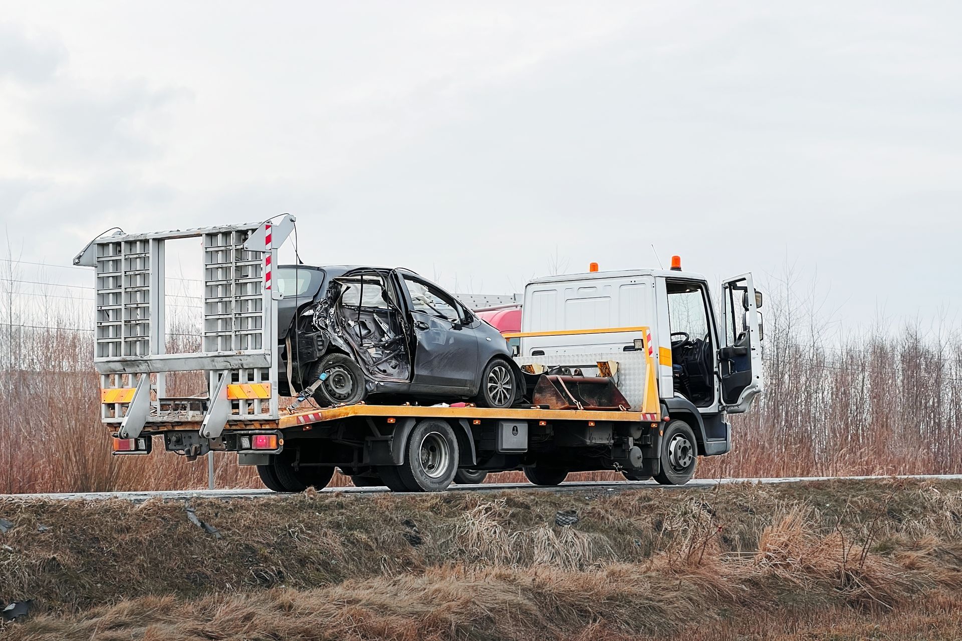 A flatbed tow truck carries a heavily damaged, burnt black car along a roadside with dry, brushy vegetation.