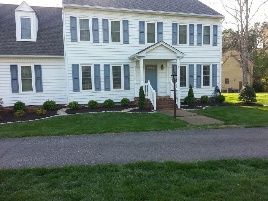 A two-story white house with blue shutters and a paved walkway leading to the front door, set against a green lawn.
